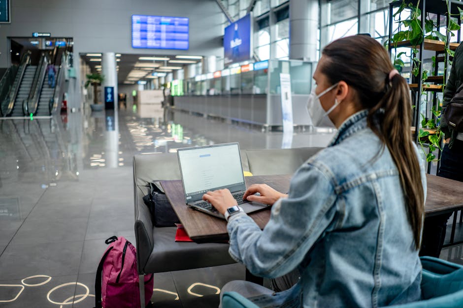 Side view of concentrated female freelancer in protective mask typing on netbook while sitting at table in modern airport terminal